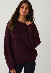 Vrouw met lang bruin haar, draagt zilveren hoepel oorbellen en een getextureerde donkerpaarse trui, terwijl ze met één hand de halslijn vasthoudt.