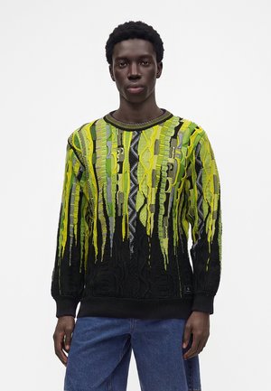 Jeune homme noir portant un pull à motifs jaune vif, vert et noir avec un jean bleu, debout devant un fond blanc uni.