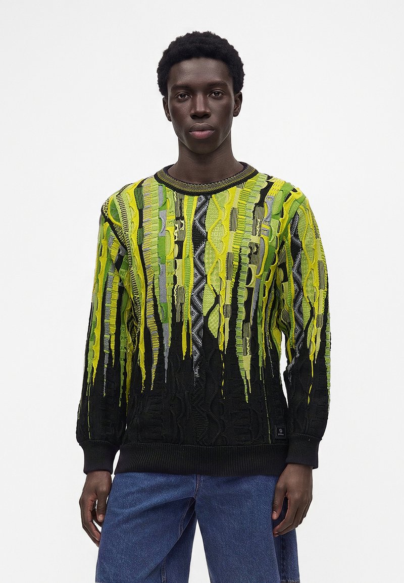 Jeune homme noir portant un pull à motifs jaune vif, vert et noir avec un jean bleu, debout devant un fond blanc uni.