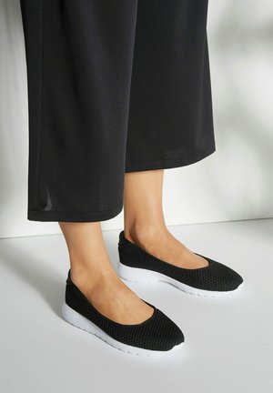 Zwarte mesh instapschoenen met een ronde neus, witte loopzool en een gestructureerde bovenzijde. Gecombineerd met losse zwarte broek.