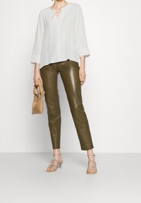 Donna che indossa una blusa bianca a maniche lunghe, pantaloni in pelle verde oliva, sandali con tacco beige e stringhe, mentre tiene in mano una piccola borsa color tan.