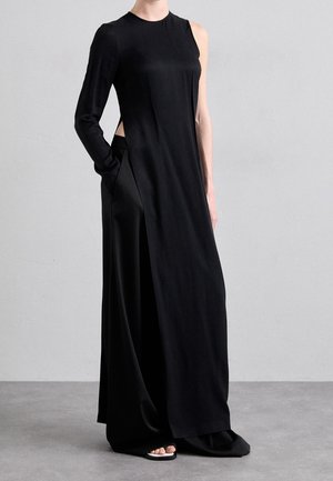 Osoba ubrana w długą czarną sukienkę z jednym długim rękawem i jedną boczną częścią bez rękawa, stojąca na szarej podłodze na tle gładkiej ściany.