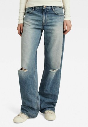 Personne portant un jean large bleu délavé avec des déchirures aux genoux et des baskets crème, les mains reposant de chaque côté.