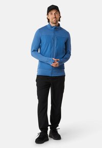Hellblaues Zip-Jackett mit gerippter Textur, ausgestattet mit einem hohen Kragen und Bambusärmelbündchen. Kombiniert mit schwarzen Hosen und Sneakers.