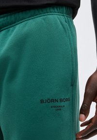 Κοντινό πλάνο των πετρόλ παντελονιών φόρμας Björn Borg με ελαστική ζώνη μέσης και μαύρο λογότυπο κοντά στην τσέπη, φορεμένα από άτομο με σκουρόχρωμο δέρμα.