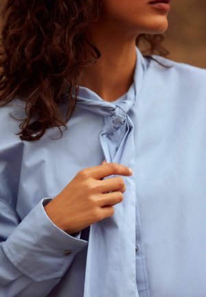 Žena s kudrnatými vlasy v světle modré blůze si upravuje uvázaný uzel u límce poblíž krku.
