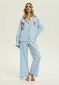 Kobieta stojąca boso, ubrana w jasnoniebieski zestaw piżam z haftem czerwonej kokardki na piersi i czerwonym obszyciem na krawędziach.