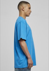 Tricou cu mânecă scurtă, lejera, de culoare albastru deschis, din bumbac, cu guler rotund și margine dreaptă, expus din lateral.