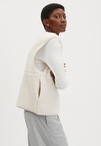 Fleeceweste in Off-White mit weicher Textur, ärmellosem Design, Seitentaschen und abgerundetem Saum. Kombiniert mit einem leichten Langarmshirt.