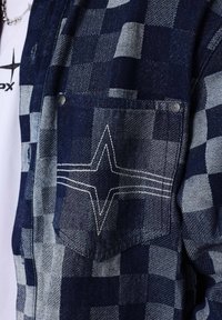 Chaqueta a rayas azul y gris con bolsillo, diseño de estrella bordada en blanco y remaches de metal, llevada sobre una camisa blanca con un logo de estrella negra.