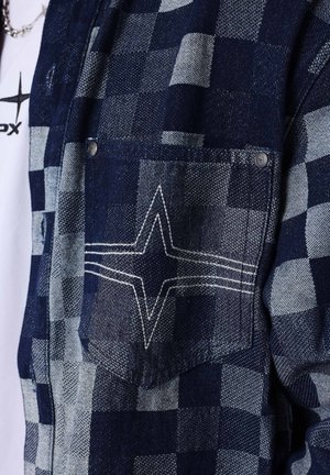Chaqueta a rayas azul y gris con bolsillo, diseño de estrella bordada en blanco y remaches de metal, llevada sobre una camisa blanca con un logo de estrella negra.