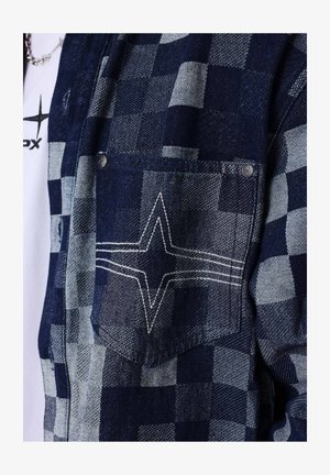 Blau- und graues kariertes Jackenetikett mit weißem gesticktem Sternmuster, Metallnieten, getragen über einem weißen Hemd mit schwarzem Sternlogo.