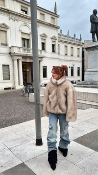 Cappotto in pelliccia sintetica color beige chiaro, oversize, con collo alto. Indossato sopra jeans cargo blu con orlo sfilacciato. Stivali neri con tacco e volant.