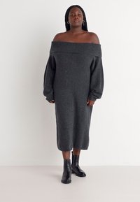 Robe grise en tricot à manches longues tombant aux épaules, longueur genoux, texture douce, portée avec des bottines noires à la cheville, design minimal, sans motifs ni accents visibles.