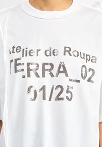 Fehér pamut póló, feltűnő, kopott szürke felirattal: "Atelier de Roupa TERRA_02 01/25." Rövid ujjú és kerek nyakú kialakítás.