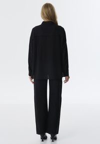 Camicia nera con bottoni, maniche lunghe, tessuto strutturato, piega sul retro e una vestibilità ampia. Abbinata a pantaloni neri a gamba larga con dettagli ricamati.