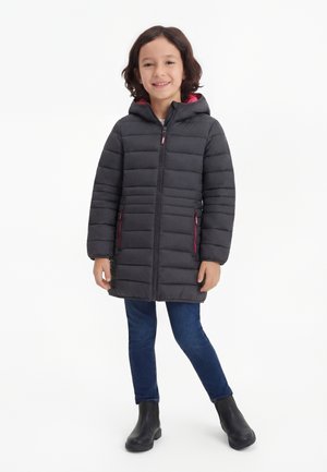 Dieťa nosiace čierny prešívaný kabát s kapucňou a ružovými zipsovými detailmi, modré džínsy a čierne topánky, stojí s jednou nohou prekríženou.