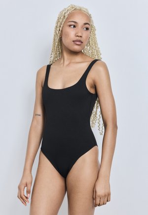 Giovane donna con capelli biondi e ricci che indossa un costume da bagno nero senza maniche, che guarda di lato su uno sfondo chiaro e uniforme.