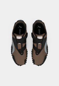 O pereche de sneakers Puma, maro și negru, cu tălpi zimțate și curele încrucișate, priviți de sus pe un fundal alb.