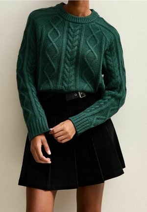 Kvinde iført en mørkegrøn kabelstrikket sweater, stoppet ned i et sort plisseret skørt med sort bælte mod en ensfarvet baggrund.