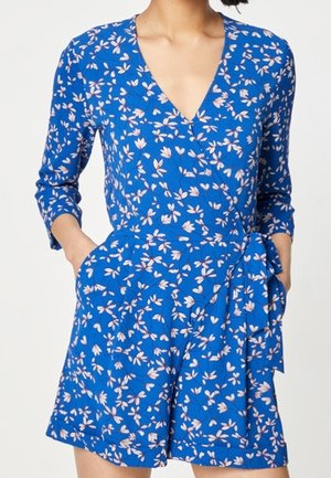 Femme portant une robe portefeuille bleue à motifs floraux avec des manches trois-quarts, un col en V, des poches latérales et une ceinture en tissu nouée à la taille.