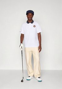 Balti polo marškinėliai su tamsiai mėlynu apykakle ir logotipu, smėlio spalvos kelnės, balti golfo batai su mėlynais akcentais, laiko golfo lazdą, dėvi tamsiai mėlyną kepurę.