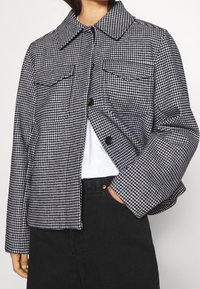 Kurtka w wzór houndstooth w czarno-białych kolorach, z kołnierzem rozłożonym, dwiema przednimi kieszeniami, zapięciem na guziki oraz luźnym krojem.