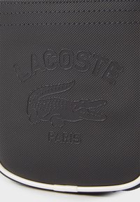 Black textured fabric wallet featuring embossed Lacoste logo and crocodile graphic, with white piping along the edges.