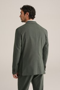 Bărbat cu păr creț și închis la culoare, purtând un costum verde măsliniu croitorit, văzut din spate pe un fundal simplu, deschis la culoare.