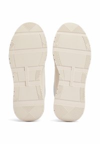 Białe podeszwy sneakersów z teksturowanymi wzorami gripowymi i podniesionymi elementami. Materiał wydaje się być gumowy o gładkim wykończeniu.