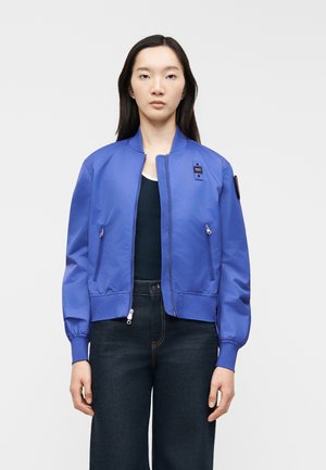 Giovane donna che indossa una giacca bomber azzurra brillante sopra una maglia scura e jeans a vita alta scuri, in piedi contro uno sfondo chiaro e uniforme.