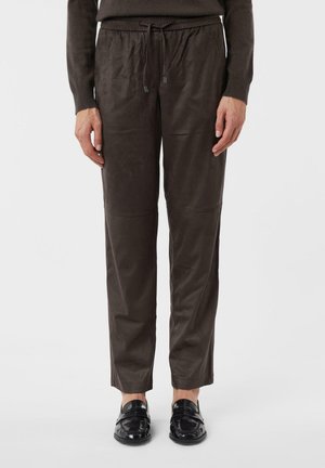 Person trägt braune Hosen mit Kordelzug und schwarze Loafers und steht vor einem weißen Hintergrund.