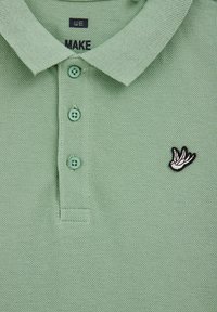 Svetlo zelena, teksturirana polo majica s pletenim ovratnikom, trima dugmadi i crno-belim izvezenim logotipom ptice na lijevoj strani prsnog koša.