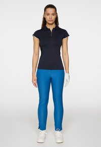 Marinblå polo med kort ärm och knäppning fram, matchad med blå tajta byxor och vita sportskor. Vit handske på vänster hand.