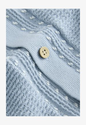 Țesătură tricotată de culoare albastră deschisă, cu un model texturat și accente albe, dotată cu un nasture rotund din lemn, decorat cu două găuri.