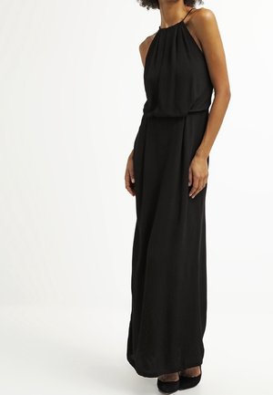 Femme portant une robe maxi noire sans manches avec un col halter, un haut plissé ample et une taille cintrée, debout devant un fond blanc uni.
