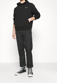 Muž v černé mikině Dickies, černých zúžených kalhotách a černobílých vysokých teniskách stojí s jednou rukou v kapse.