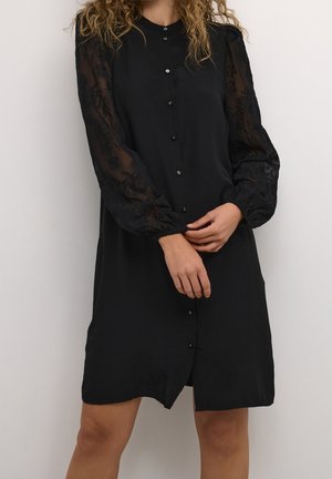 Femme portant une robe noire boutonnée avec des manches longues transparentes brodées, debout contre un fond clair uni.