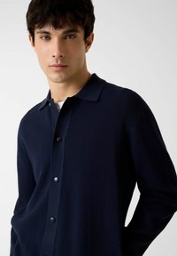 Camicia a maglia blu navy con bottoni, colletto, tessuto strutturato e grandi bottoni. Il design è aderente con una sottile lucentezza.