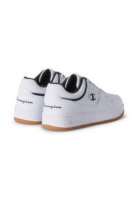 Para białych sneakersów Champion z czarnymi detalami, gumowymi podeszwami, logo Champion na bokach, piętach i środkowych częściach podeszwy, zaprezentowane z tylnego kąta.
