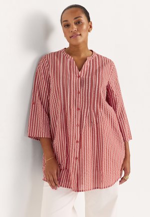 Vrouw draagt een rood-wit gestreept blouse met verticale strepen, knopen en driekwart mouwen, en witte broek, staand tegen een witte muur.