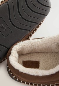 Bruna mockasiner i mocka med en mjuk krämfärgad foder, som har en texturerad svart gummisula med mönster och dekorativt sömnad.