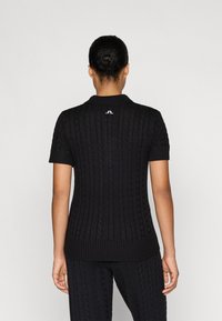 Polo de manga corta negro con textura de punto de cable, cuello y dobladillo acanalados, y un pequeño detalle de logo en la parte posterior.