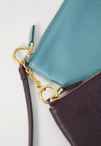 Duos tekstūruotų odinių maišelių, vienas melsvai žalias, o kitas tamsiai violetinis, sujungtų auksiniu grandinėle ir užtrauktukų traukikliais ant balto fono.