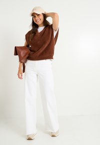 Un gilet en tricot marron et duveteux sur un t-shirt blanc, associé à un jean blanc à jambes larges et des baskets beiges. Un sac à main marron et une casquette beige complètent le look.