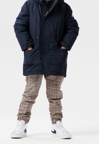 Veste doudoune marine avec capuche, fermeture éclair et boutons ; pantalons à carreaux en beige, rouge et vert ; baskets blanches avec accents noirs.