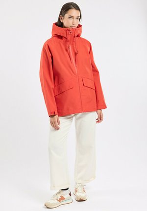 Femme portant une veste à capuche orange vif, un pantalon large blanc et des baskets beiges, debout devant un fond blanc uni.