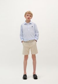 Camicia a maniche lunghe azzurro chiaro con colletto abbottonato e texture a motivi, abbinata a pantaloni corti beige e mocassini neri.