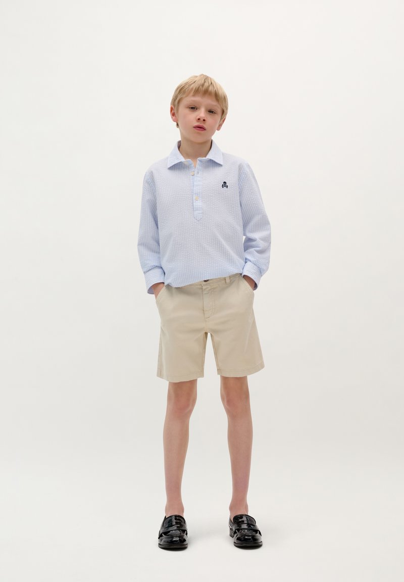 Camicia a maniche lunghe azzurro chiaro con colletto abbottonato e texture a motivi, abbinata a pantaloni corti beige e mocassini neri.