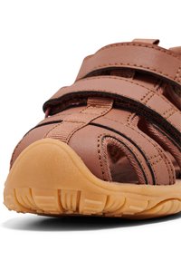 Brązowy sandał zamknięty z regulowanymi paskami i teksturowaną jasnobrązową gumową podeszwą, zaprojektowany z myślą o komforcie i przepuszczalności powietrza.